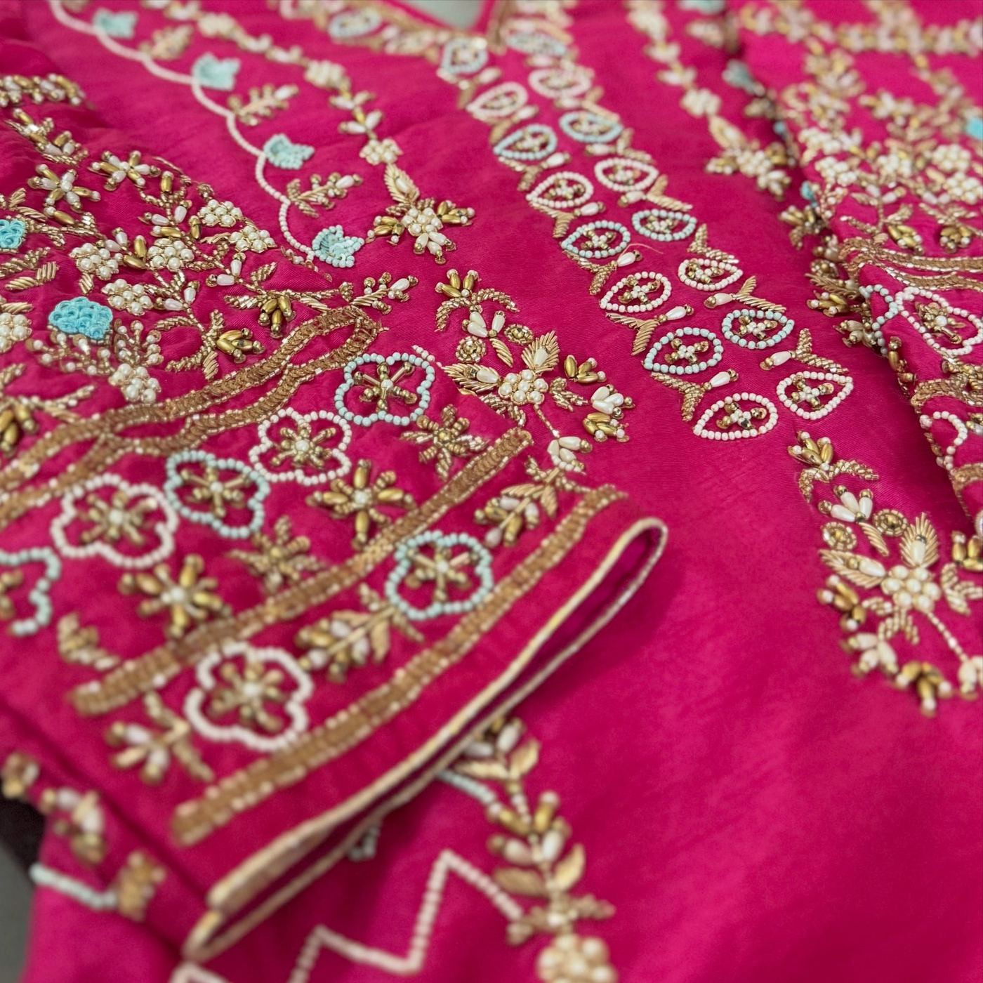 Close-up of fuchsia-pink bridal fabric with dense gold gota, pearl-bead, and turquoise floral embroidery.
