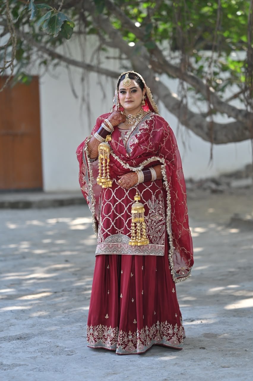 Bride standing in a courtyard wearing a maroon bridal sharara set with a silver lattice-pattern gota embroidered top and skirt, sheer maroon dupatta, and gold kaleere.