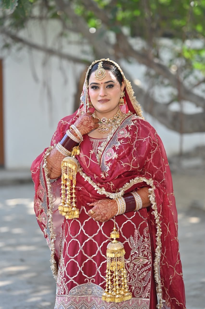 Close portrait of the bride in the maroon lattice gota sharara set, dupatta draped over her head, gold maang tikka and layered necklace, gold kaleere in hand.
