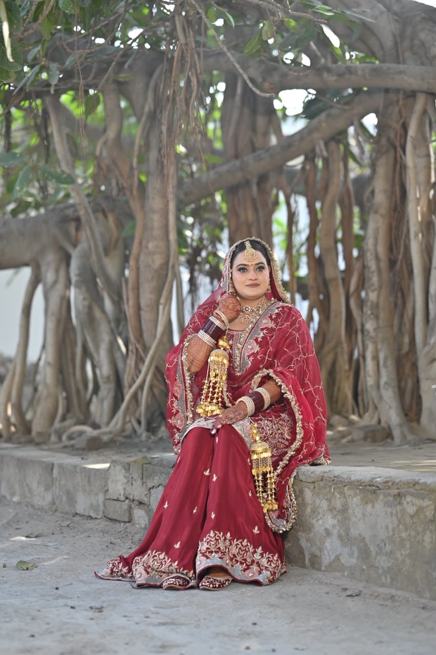 Bride seated on stone steps under a banyan tree, wearing a maroon lehenga and dupatta with delicate gold vine embroidery, gold jewelry, and gold kaleere.