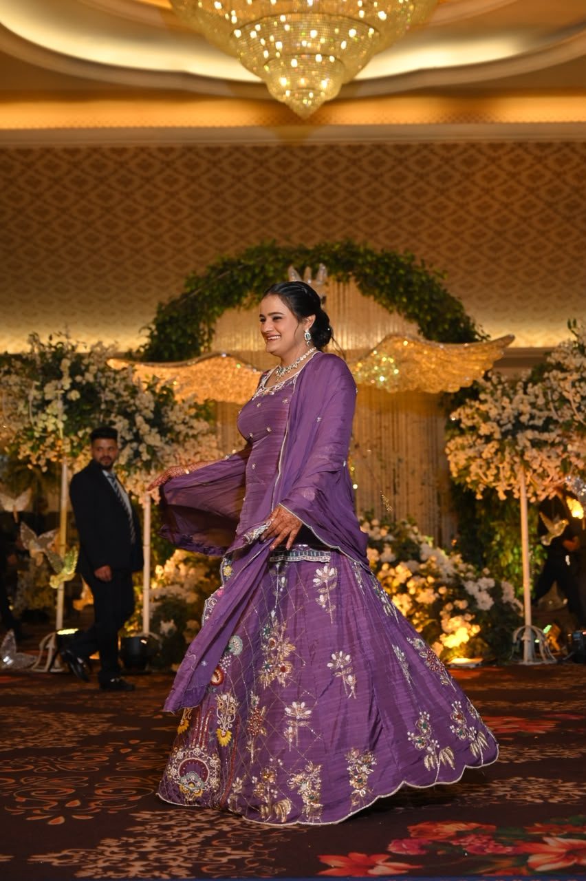 Woman twirling in a lavender-purple lehenga at a wedding reception, the skirt embroidered with gold and silver floral and paisley motifs, dupatta draped over one shoulder.