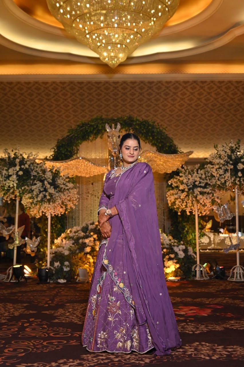Woman at a wedding reception wearing a deep-purple lehenga with a dupatta draped saree-style over one shoulder, gold floral embroidery along the skirt hem.
