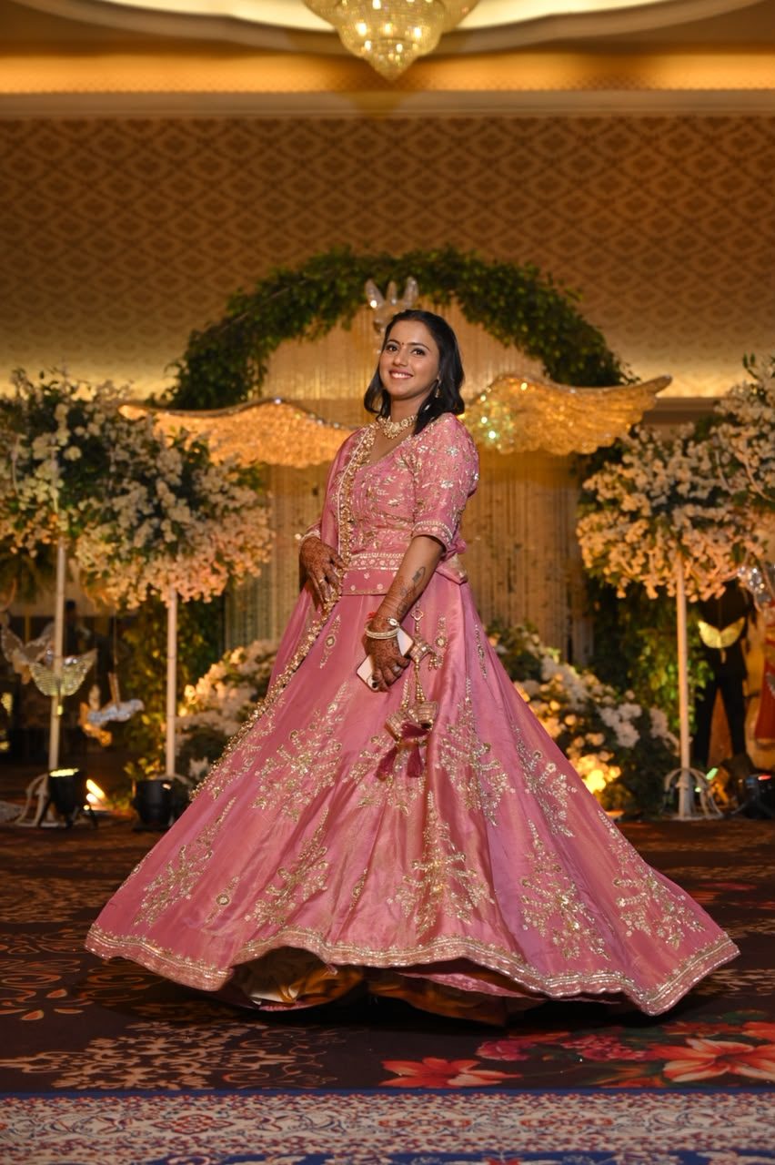 Front view of the rose-pink floral-embroidered lehenga mid-twirl at a wedding reception, gold embroidered bodice and skirt border, dupatta draped over the shoulder.