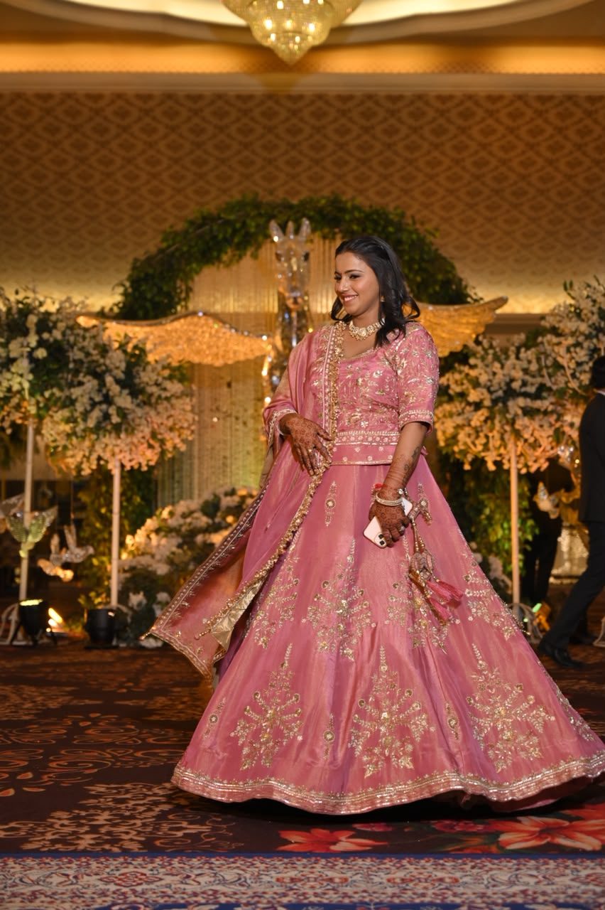 Woman twirling in a rose-pink lehenga at a wedding reception, the skirt densely embroidered with gold floral and paisley motifs, dupatta flowing to the side.