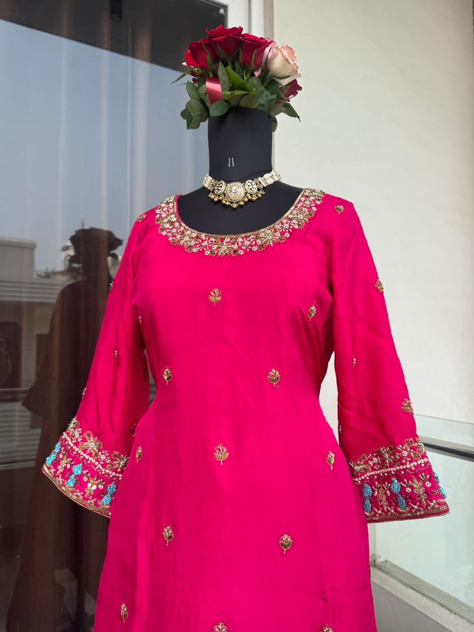 Close front view of a hot-pink suit with a gold and pearl floral-cluster embroidered neckline and small floral booti scattered across the bodice.